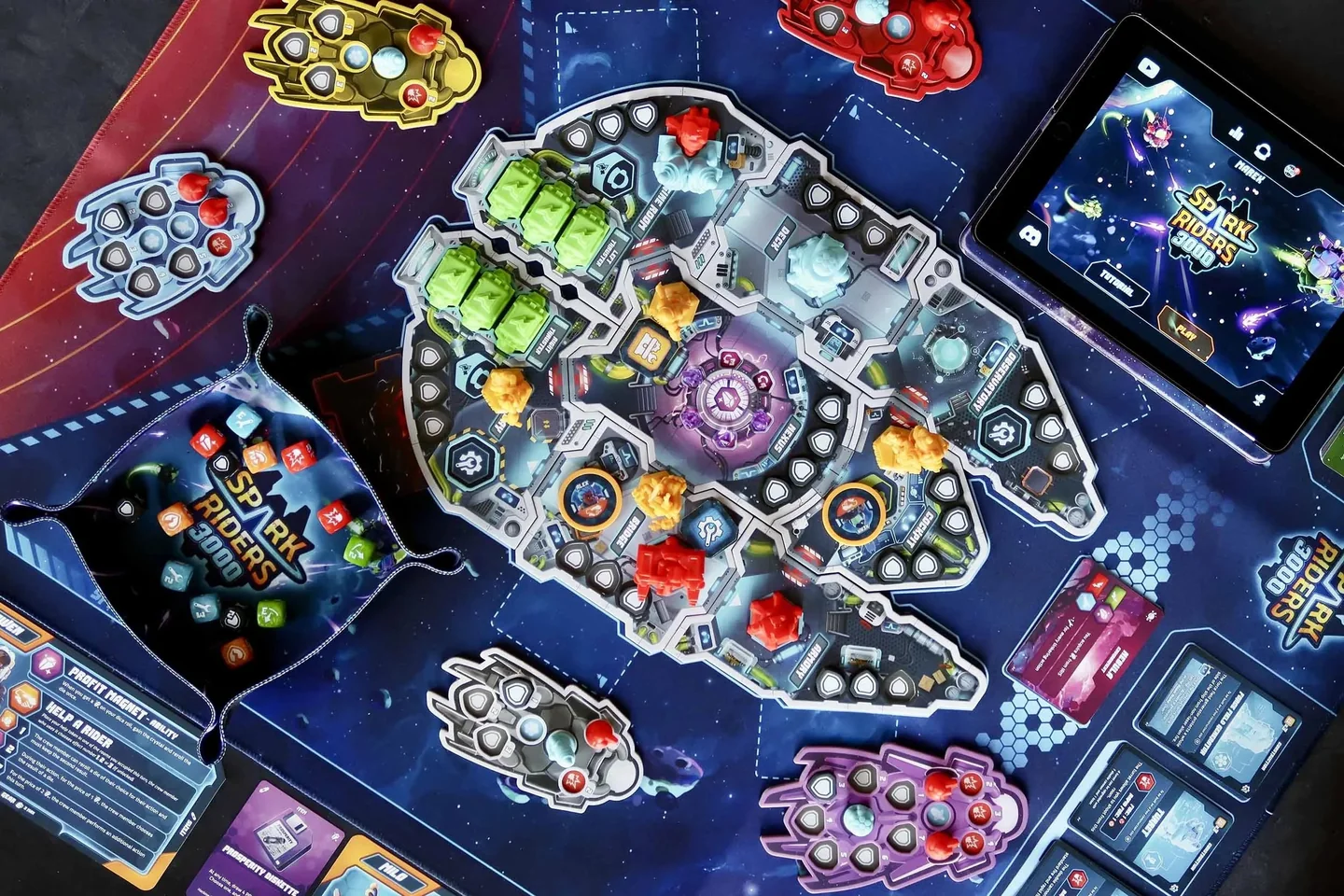 overhead view of the Spark Riders 3000 spaceship board, rider panels, cards and dice, with a tablet at upper right.